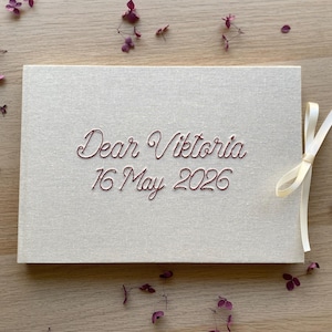 May include: A rectangular, light beige fabric-covered album featuring the embroidered text "Dear Viktoria 16 May 2026" in a dark red script font. A cream-colored ribbon is tied in a bow on the right side of the album cover.