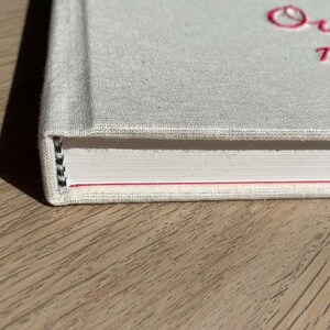May include: Close-up of a hardcover book with a light gray linen cover. The book's title, "Our 72 Seasons," is embroidered in pink thread. The spine shows the bound pages with a black and white thread.