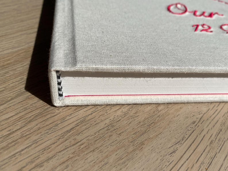 May include: Close-up of a hardcover book with a light gray linen cover. The book's title, "Our 72 Seasons," is embroidered in pink thread. The spine shows the bound pages with a black and white thread.