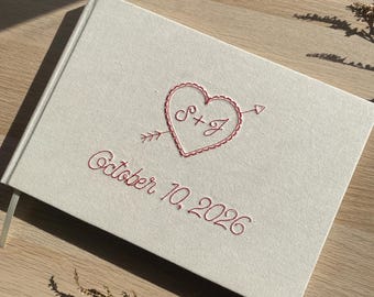 May include: A rectangular off-white fabric-covered guest book featuring red embroidered details. The cover displays a heart pierced by an arrow with the initials S+J inside, and the text October 10, 2026, stitched in red thread below the heart.