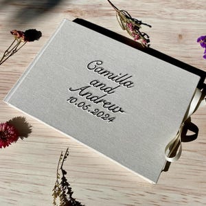 Może przedstawiać: Biały gościnny album z lnu z białą wstążką satynową. Okładka jest tłoczona tekstem "Camilla i Andrew 10.05.2024".