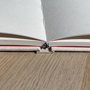 May include: An open notebook with blank white pages. The binding is visible, showing a red stripe and a textured, light-colored cover. The notebook is on a wooden surface.