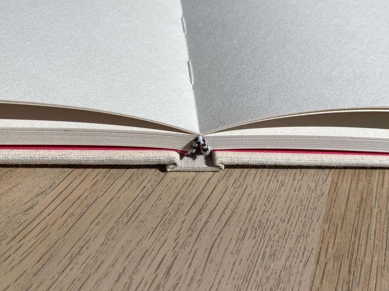 May include: An open notebook with blank white pages. The binding is visible, showing a red stripe and a textured, light-colored cover. The notebook is on a wooden surface.