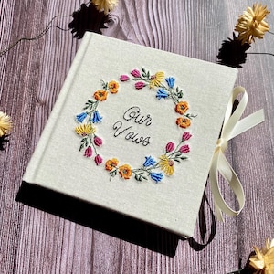 May include: A white linen guest book with a wreath of embroidered flowers in shades of yellow, blue, pink, and green. The text "Our Vows" is embroidered in the center of the wreath.