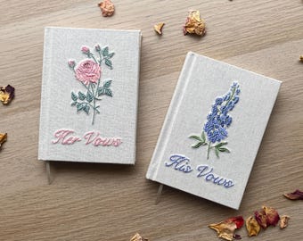 May include: Two light gray fabric-covered vow books with embroidered floral designs. One book features a pink rose and the text "Her Vows" in pink thread. The other book features a blue lupine flower and the text "His Vows" in blue thread.