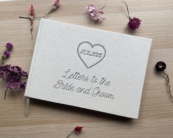 May include: A light-colored wedding guest book with the text "Letters to the Bride and Groom" embroidered in black. The cover features a heart with the date 27.12.2025. Dried flowers in shades of purple and pink are scattered around the book.