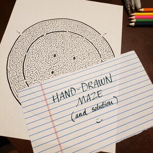 Könnte beinhalten: Handgezeichnetes, kreisförmiges Labyrinth in Schwarzweiß auf weißem Papier. Eine Notiz mit der Aufschrift "HAND-DRAWN MAZE (and solution)" und einem Smiley. Das Labyrinth hat ein komplexes Muster. Buntstifte und Stifte sind sichtbar.