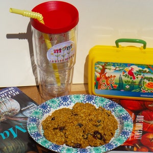 Pu&ograve; includere: Primo piano di un piatto di biscotti all'avena, uvetta e mirtilli rossi. I biscotti sono su un piatto di carta con motivo blu e bianco. C'&egrave; una scatola da pranzo gialla con una scimmia, un tucano e una tartaruga disegnati su di essa. C'&egrave; un bicchiere trasparente con un coperchio rosso e una cannuccia gialla. Il bicchiere ha un adesivo che dice "Mamma" su di esso. Ci sono due libri sullo sfondo, uno con una zucca sulla copertina e l'altro con un titolo che dice "Riviste".