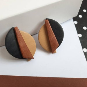Puede incluir: Un par de pendientes geométricos. Cada pendiente presenta un semicírculo negro, un semicírculo beige y un triángulo de madera marrón. Los pendientes están sobre un fondo blanco con un patrón de lunares blancos y negros.