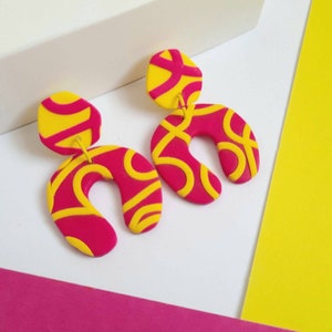 May include: A pair of vibrant, handmade earrings. The earrings feature a bright pink, U-shaped design with yellow, swirled accents. The top portion is a yellow oval shape. The earrings are a bold statement piece.