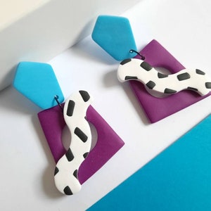 Könnte beinhalten: Ein Paar abstrakte Ohrringe mit geometrischem Design. Jeder Ohrring besteht aus einem hellblauen, fünfeckigen Oberteil, einem lila Quadrat mit Loch und einer weißen, wellenförmigen Form mit schwarzen Punkten. Vor weißem und blauem Hintergrund.