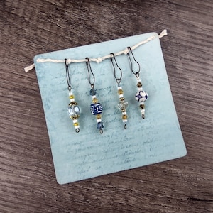 May include: Set of four blue and white beaded stitch markers with gold accents. The markers are attached to a blue card with white writing.