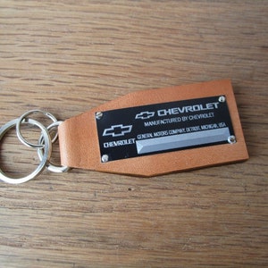 Puede incluir: Un llavero de cuero marrón con una etiqueta de metal plateado que dice "CHEVROLET MANUFACTURED BY CHEVROLET GENERAL MOTORS COMPANY, DETROIT, MICHIGAN, USA".