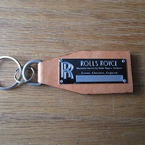 以下が含まれることがあります： 「ROLLS ROYCE」と「Manufactured by Rolls-Royce Limited Crewe, Cheshire, England」と書かれた銀色の金属タグが付いた茶色の革製のキーホルダーです。