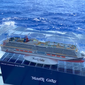 May include: A white and blue model cruise ship with the words "Mardi Gras" and "Carnival" on the side. The model is sitting on a blue box with the words "Mardi Gras" on it. The model is in front of a blue ocean.