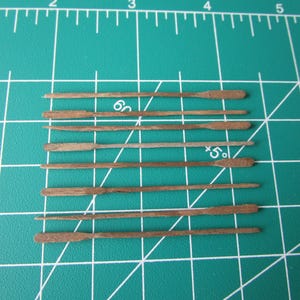 May include: Eight wooden oars, all the same size and shape, are lined up on a green gridded surface. The oars are brown and have a slight curve at the end.