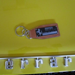 Puede incluir: Un llavero Ferrari de cuero marrón con un llavero plateado. El llavero tiene una placa negra con el logotipo de Ferrari y texto. El fondo es una superficie de color amarillo brillante, con el logotipo cromado de Ferrari visible.