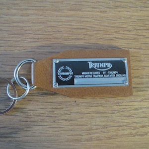 Op de afbeelding: Een bruin leren sleutelhanger met een zilveren sleutelring. De sleutelhanger heeft een rechthoekige metalen plaat met het woord "TRIUMPH" en de tekst "MANUFACTURED BY TRIUMPH TRIUMPH MOTOR COMPANY, COVENTRY, ENGLAND".