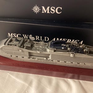 May include: A silver-toned metal model of the MSC World America cruise ship, featuring detailed deck structures and a dark blue funnel. The model is mounted on a rectangular maroon base and is positioned in front of a black box labeled MSC WORLD AMERICA.