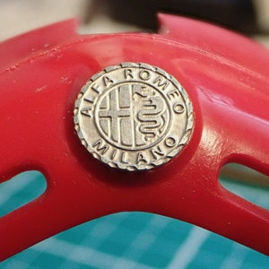 Puede incluir: Primer plano de un emblema Alfa Romeo Milano plateado con un logotipo de serpiente sobre un fondo rojo.