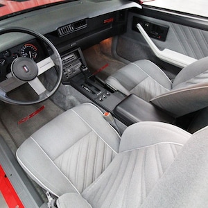 May include: The interior of a red car with grey cloth seats and a black steering wheel. The car has a black dashboard with a radio and other controls. The car has a manual transmission.