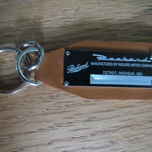 May include: A brown leather keychain with a silver key ring and a black metal tag that reads "Packard, Manufactured by Packard Motor Company, Detroit, Michigan, USA".