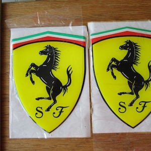 May include: Two yellow and black Ferrari car emblems with a black horse rearing up on its hind legs. The emblems are on a white background with a green, white, and red stripe at the top.