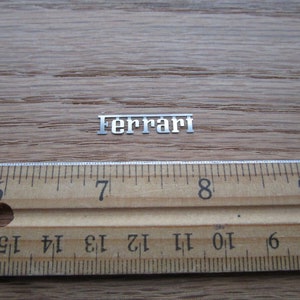 Puede incluir: Un logotipo de Ferrari de metal plateado sobre una superficie de madera con una regla para la escala. El logotipo mide aproximadamente 2,5 cm de largo.