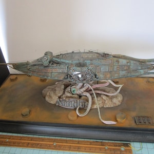 May include: A detailed model of the submarine The Nautilus, featuring a weathered teal and copper finish, mounted above a sculpted octopus on a rectangular base. A wooden ruler is visible at the bottom, showing a length of 12 inches.