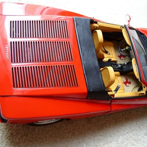 以下が含まれることがあります： 黒の幌とベージュのインテリアを備えた赤いフェラーリ コンバーチブル モデルカー。車は、黒いグリルと側面に印刷された「フェラーリ」という文字を持つ詳細なエンジンルームを備えています。