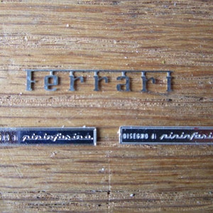 May include: Close-up of three silver metal plates with the words "Ferrari", "Disegno di Pininfarina", and "Disegno di Pininfarina" engraved on them.