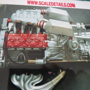 Könnte beinhalten: Nahaufnahme eines detaillierten Modells eines silbernen Motors mit roten Kabeln und einem schwarzen Schlauch. Der Motor hat einen silbernen Ansaugkrümmer und einen silbernen Auspuffkrümmer. Der Motor ist auf einem silbernen Sockel montiert. Das Bild enthält die Website-Adresse www.scaledetails.com.