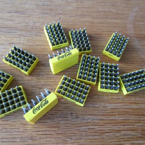 May include: Multiple miniature yellow cardboard boxes filled with tiny gray bottles. Each box has the Coca-Cola logo printed on the front.
