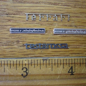 May include: Close-up of three silver metal plates with the words "Ferrari", "Disegno di Pininfarina", and "Testarossa" on a wooden surface.