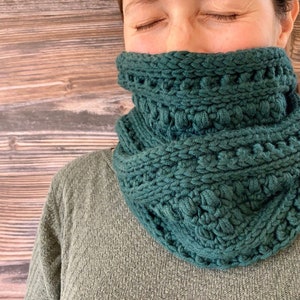 May include: A dark green knitted cowl neck scarf with a textured pattern. The scarf is folded over and worn around the neck.