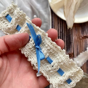 May include: A white crocheted garter with a blue satin ribbon bow. The garter is delicate and intricate, with a lacy design.