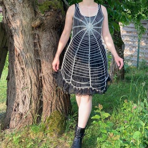 Może przedstawiać: Czarna sukienka z białym wzorem pajęczyny. Sukienka bez rękawów ma warstwową spódnicę z koronkowym wykończeniem. Osoba ma na sobie czarne buty i stoi w pobliżu drzewa.