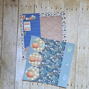 May include: Three paper circles with a checkered pattern and a pumpkin illustration on each. The circles are on a blue background with a floral pattern. The text on the circles reads "Happy Fall", "Cozy Days", and "Thankful".