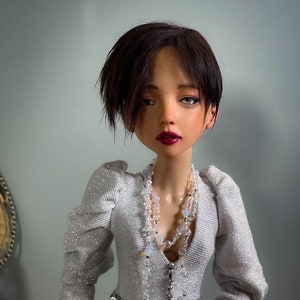 May include: A doll wearing a silver sequined dress with a silver sparkly top and a pearl necklace. The doll has short black hair and red lipstick.