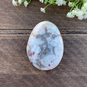 May include: A white and gray polished stone with red and pink inclusions. The stone is shaped like an egg and has a smooth, polished surface.