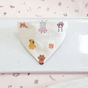 K&ouml;nnte beinhalten: Ein wei&szlig;es dreieckiges Bandana mit einem Katzenkopf, Pfotenabdr&uuml;cken und anderen Tiermotiven. Das Bandana liegt auf einer wei&szlig;en Oberfl&auml;che.