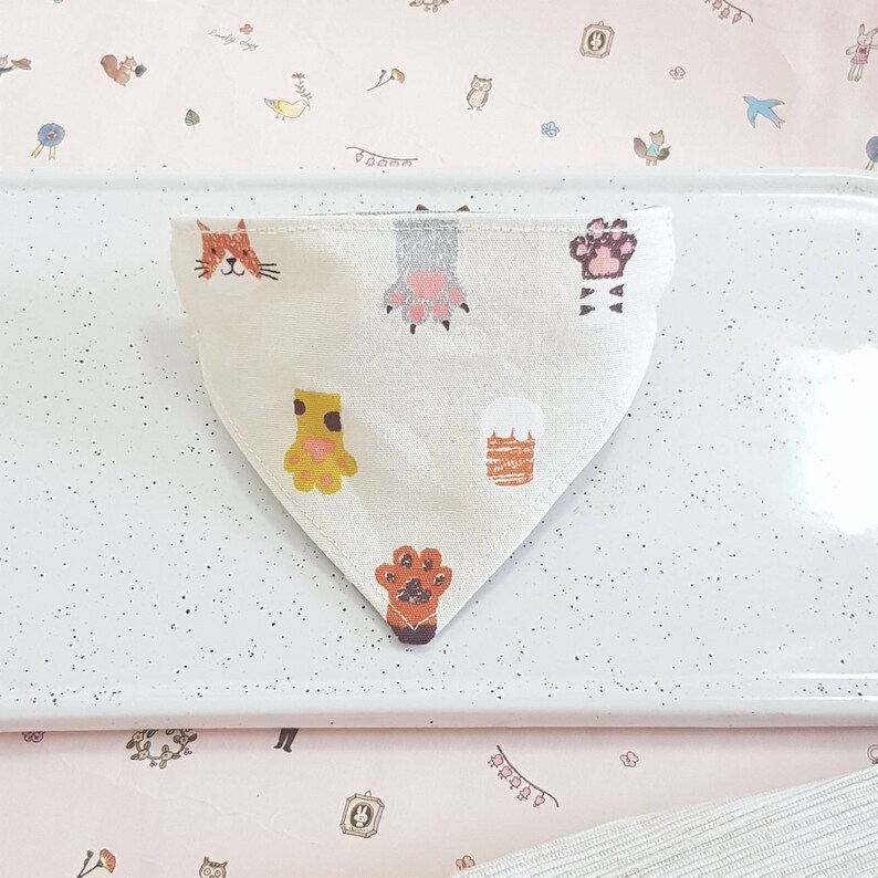 K&ouml;nnte beinhalten: Ein wei&szlig;es dreieckiges Bandana mit einem Katzenkopf, Pfotenabdr&uuml;cken und anderen Tiermotiven. Das Bandana liegt auf einer wei&szlig;en Oberfl&auml;che.