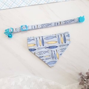 May include: A light blue cat collar with a bell and a matching light blue bandana with a pattern of yellow and blue fish.