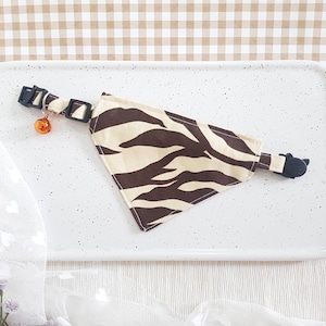 May include: A brown and beige zebra print bandana with a black adjustable collar and a small orange bell.