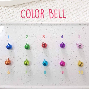 May include: Ten small jingle bells in different colors, numbered 1 through 10. The bells are arranged in two rows of five on a white surface with black speckles. The text "COLOR BELL" is written in red at the top of the image.
