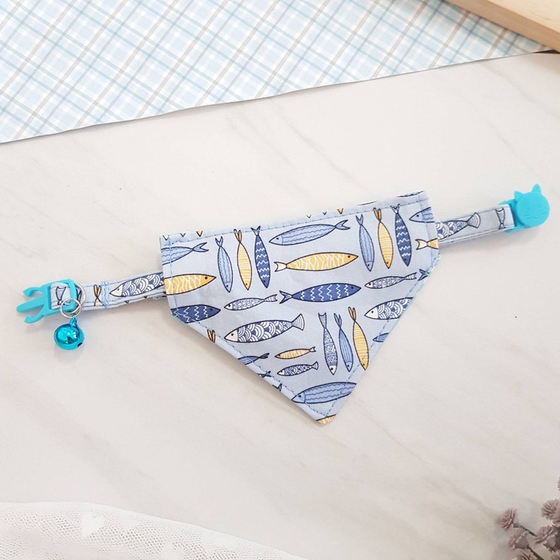 May include: A light blue cat collar with a bell and a blue fish pattern bandana. The bandana has a blue and yellow fish pattern.