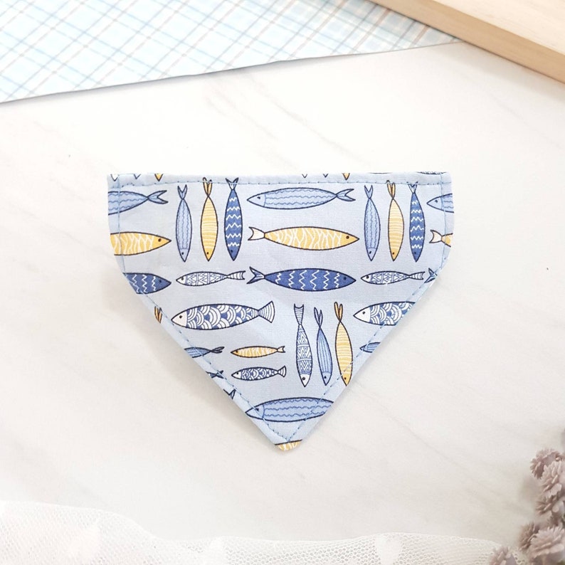 May include: A light blue bandana with a pattern of yellow and blue fish. The bandana is triangular in shape and has a white background.
