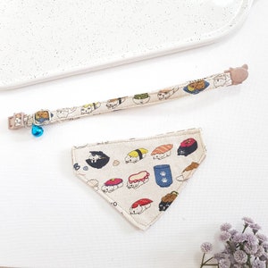 May include: A beige cat collar with a bell and a matching beige bandana with a sushi print. The bandana is triangular and features various sushi rolls with cartoon cats on them.