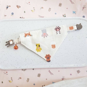 K&ouml;nnte beinhalten: Ein wei&szlig;es Baumwollbandana mit einem Katzpfoten-Design. Das Bandana hat ein braunes Katzen Gesicht, Pfotenabdr&uuml;cke und einen Katzenschwanz. Das Bandana ist an einem braunen katzenf&ouml;rmigen Clip mit einer Glocke befestigt.