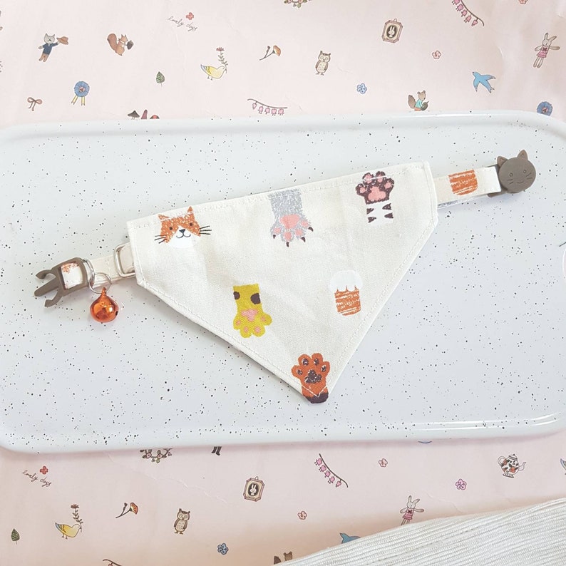 K&ouml;nnte beinhalten: Ein wei&szlig;es Baumwollbandana mit einem Katzpfoten-Design. Das Bandana hat ein braunes Katzen Gesicht, Pfotenabdr&uuml;cke und einen Katzenschwanz. Das Bandana ist an einem braunen katzenf&ouml;rmigen Clip mit einer Glocke befestigt.